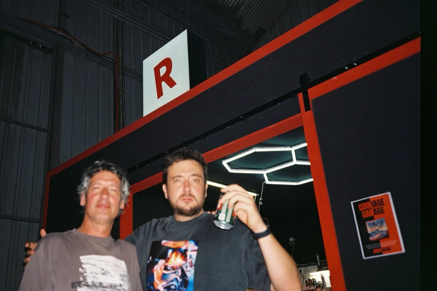 Two friends at the garage entrance under the red R sign, one raising a can