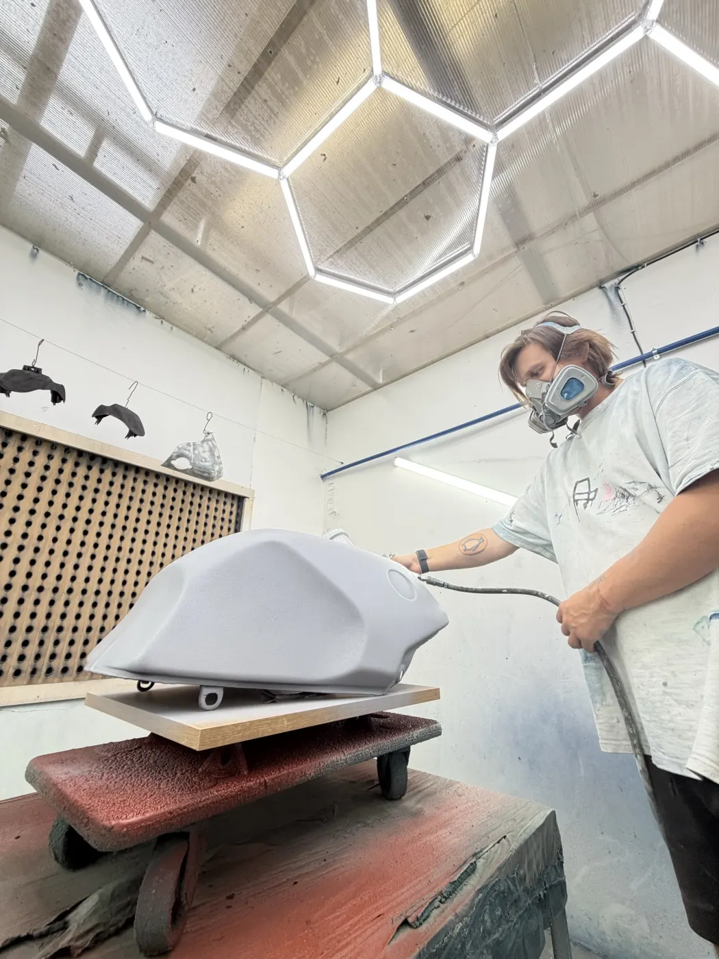 A rider in a respirator spraying primer on a motorcycle fuel tank in the paint booth